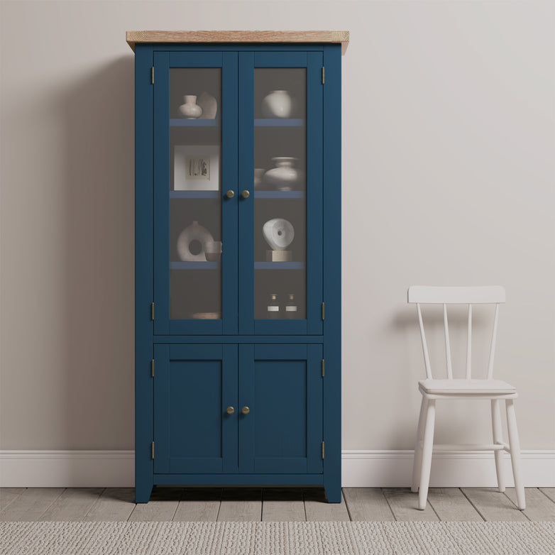 The Burford Display Cabinet in Navy Blue, featuring glass doors that beautifully display pottery, stands elegantly beside a white chair against a light-colored wall, adding a touch of sophistication and style.