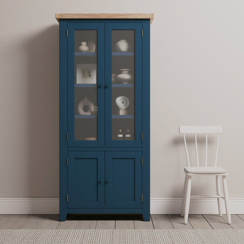 The Burford Display Cabinet in Navy Blue is positioned adjacent to a white wooden chair on a light grey rug, elegantly embellished with decorative items that accentuate its charm.