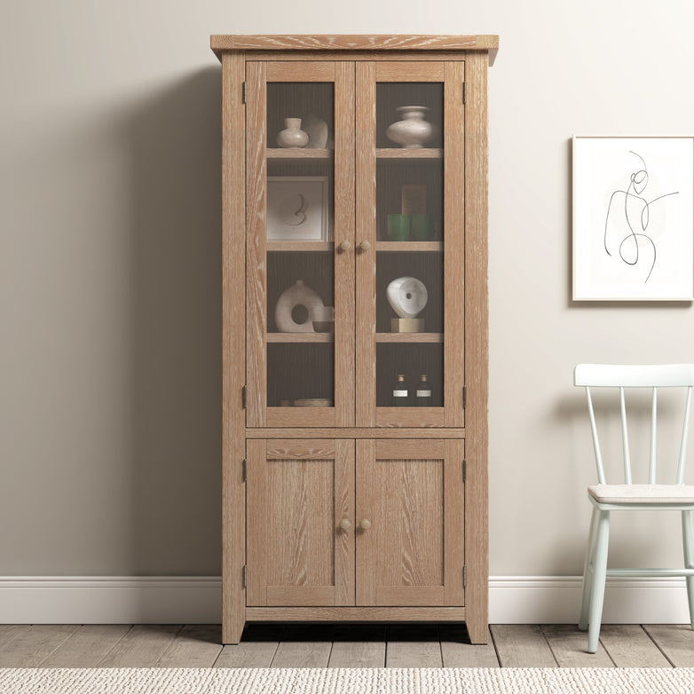 The Burford Display Cabinet in Natural Oak, featuring glass-panelled doors, beautifully showcases decorative items inside. It is positioned beside a white chair and complemented by abstract wall art.