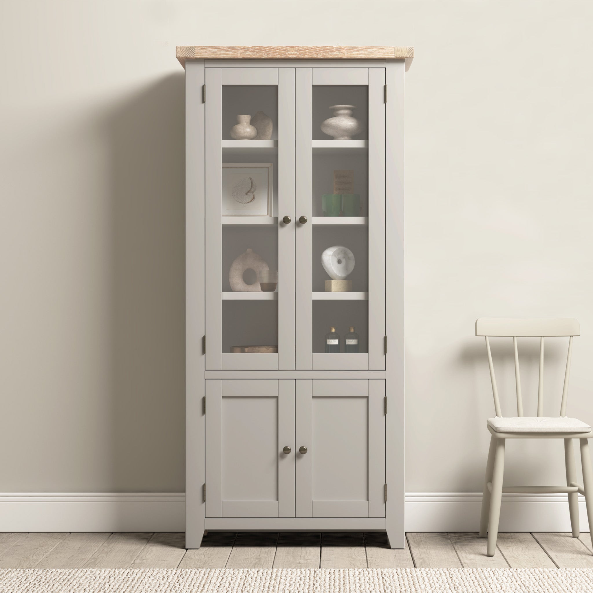 The Burford Display Cabinet in Pebble Grey, with its elegant glass-panelled doors, beautifully showcases decor items. It's complemented by a simple white chair set against a light-colored wall, creating a charming contrast. The subtle hints of natural oak introduce warmth and character into the space.