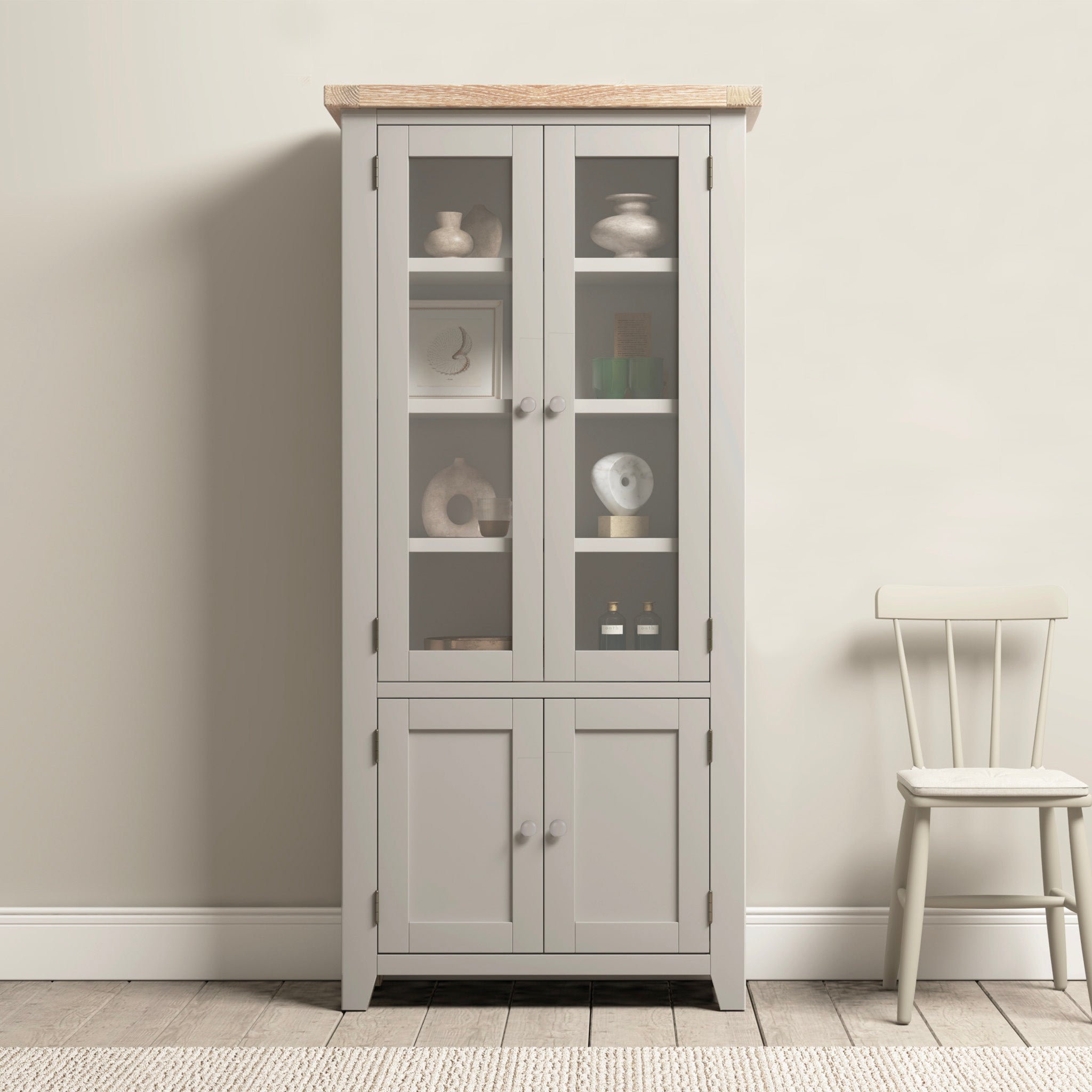 The Burford Display Cabinet in Pebble Grey, featuring glass-panelled doors, elegantly showcases decor items next to a simple wooden chair against a light wall.