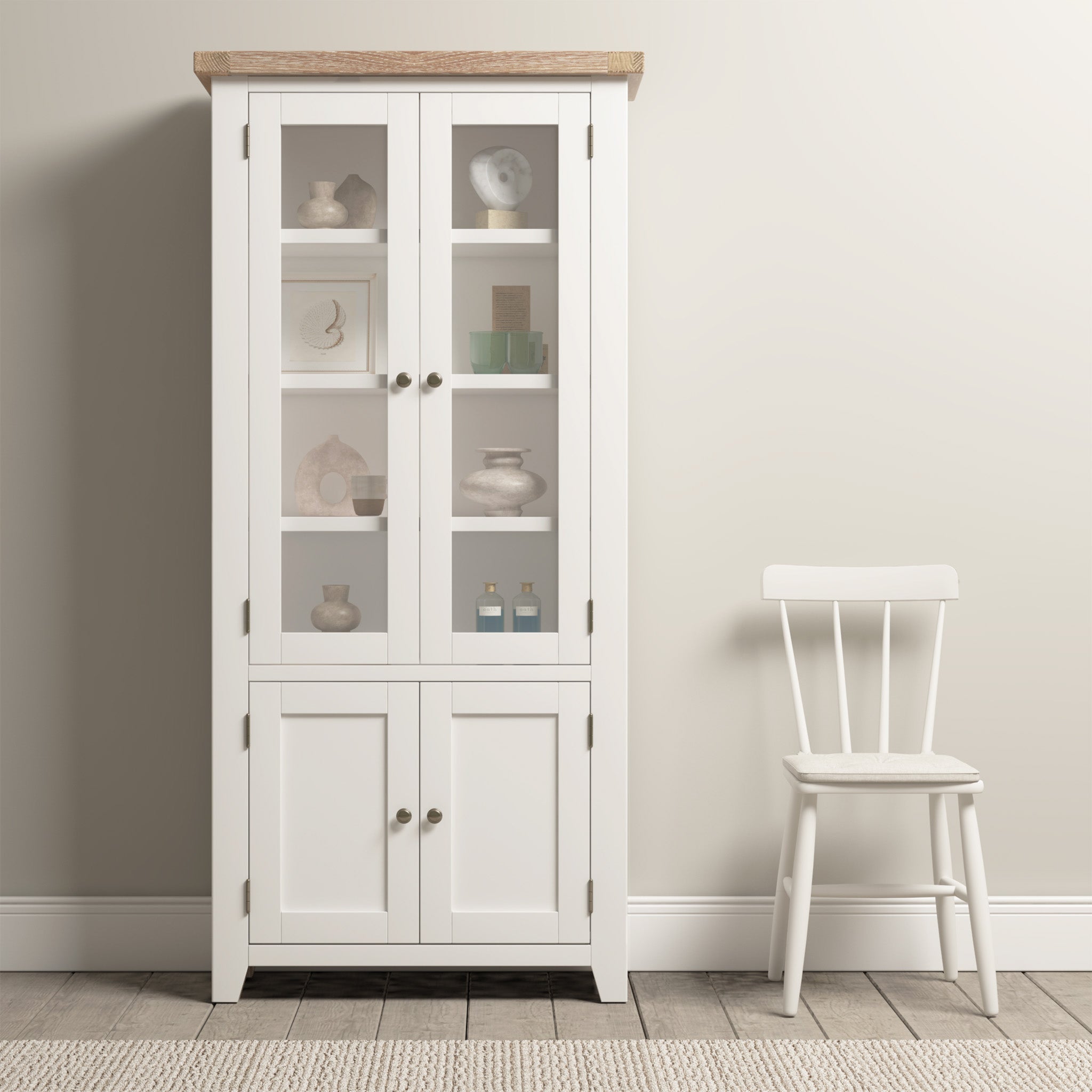 A Burford Display Cabinet in Warm White with glass-panelled doors showcases decor items beside a white wooden chair on a neutral-toned floor.