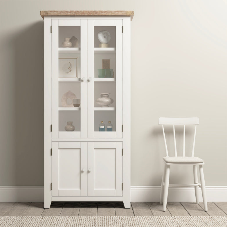 The Burford Display Cabinet in Warm White, featuring glass-panelled doors, showcases decor next to a simple white chair, all resting on a natural oak wooden floor against a beige wall.