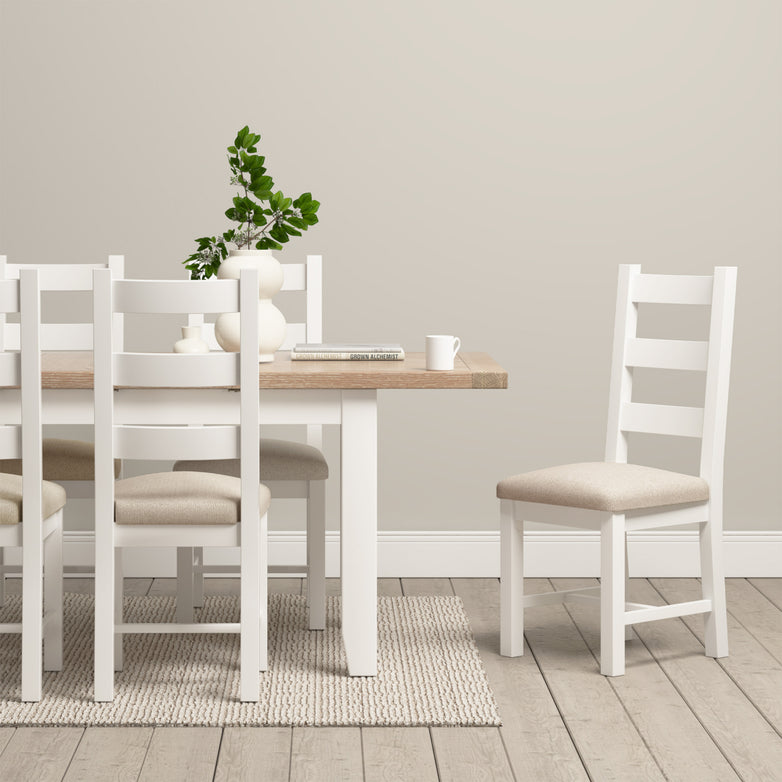 A minimalist dining room showcasing contemporary elegance features a Burford 4-6 Seater Extending Dining Table in warm white, complemented by white chairs, a vase of leaves, books, and a cup on a woven rug.