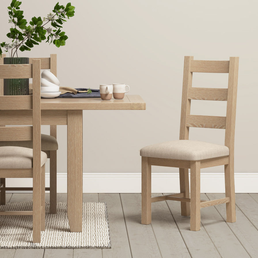 A dining table with two sets of Burford Ladderback Dining Chairs featuring fabric seats in natural oak enhances a vase with greenery and two mugs on a neutral-toned carpet, all exemplifying the elegance of natural oak in a sophisticated whitewash finish.