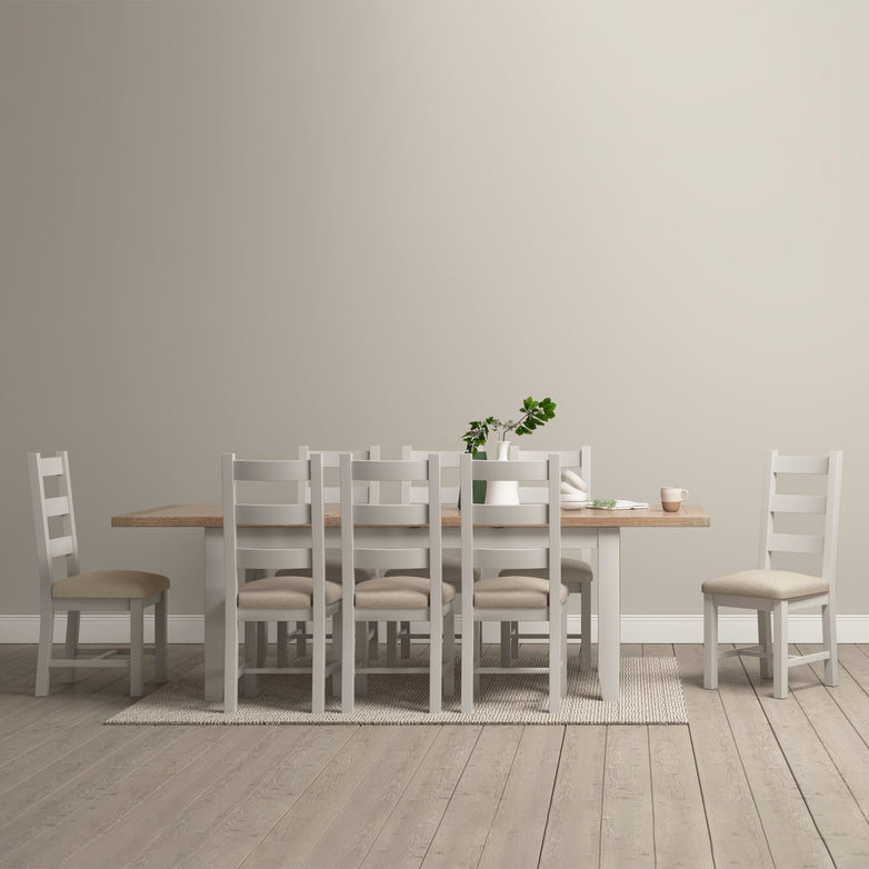 A minimalist dining room showcases a Burford 6-10 Seater Extending Dining Table in Pebble Grey, enhanced by a matching rug and surrounded by eight chairs. Natural oak finishes and potted plants complete the serene look.