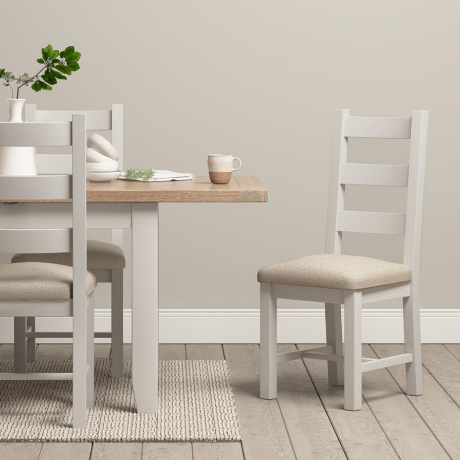 Modern dining space showcasing Pebble Grey Burford Ladderback Dining Chairs with fabric seats, complemented by a natural oak wooden table and a white vase filled with greenery.