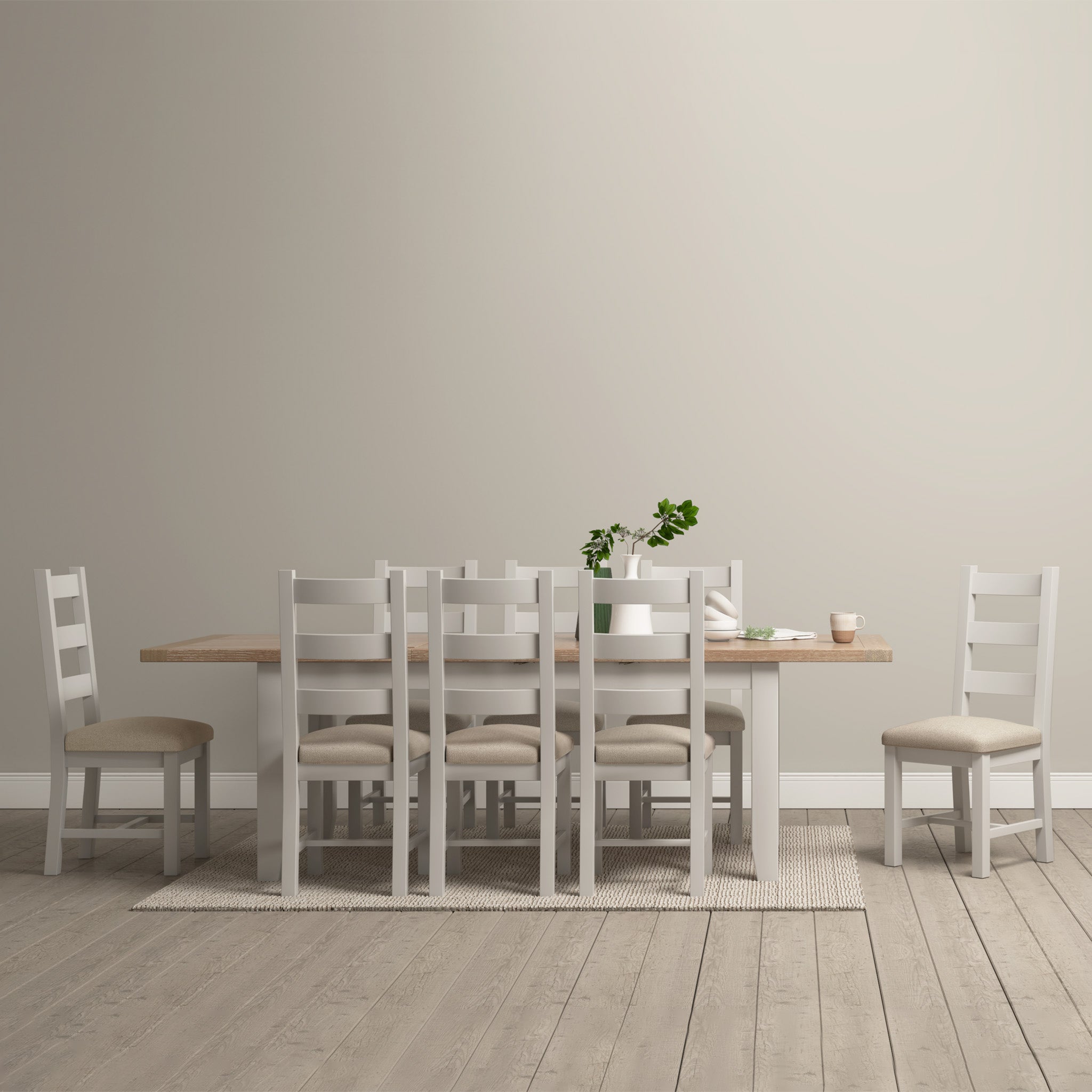 A minimalist dining room showcasing the Burford 8 Seat Extending Dining Set in Pebble Grey. The wooden table, accompanied by six chairs and decorated with plants, stands out against a backdrop of a subtle pebble grey wall.