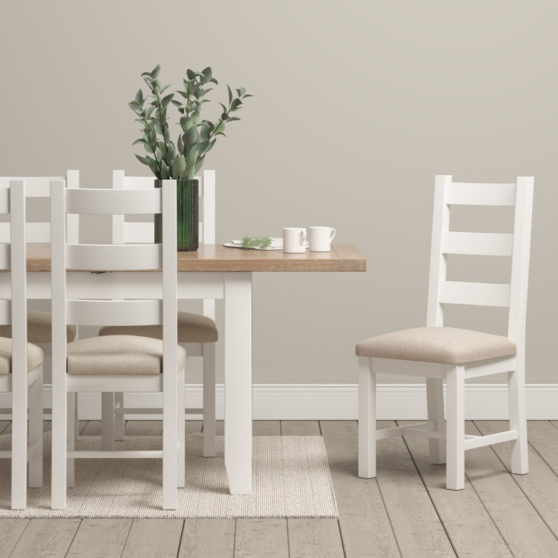 White wooden dining table paired with a set of two Burford Ladderback Dining Chairs with fabric seats in Warm White, accompanied by a plant and cups on top, set on a light natural oak floor.