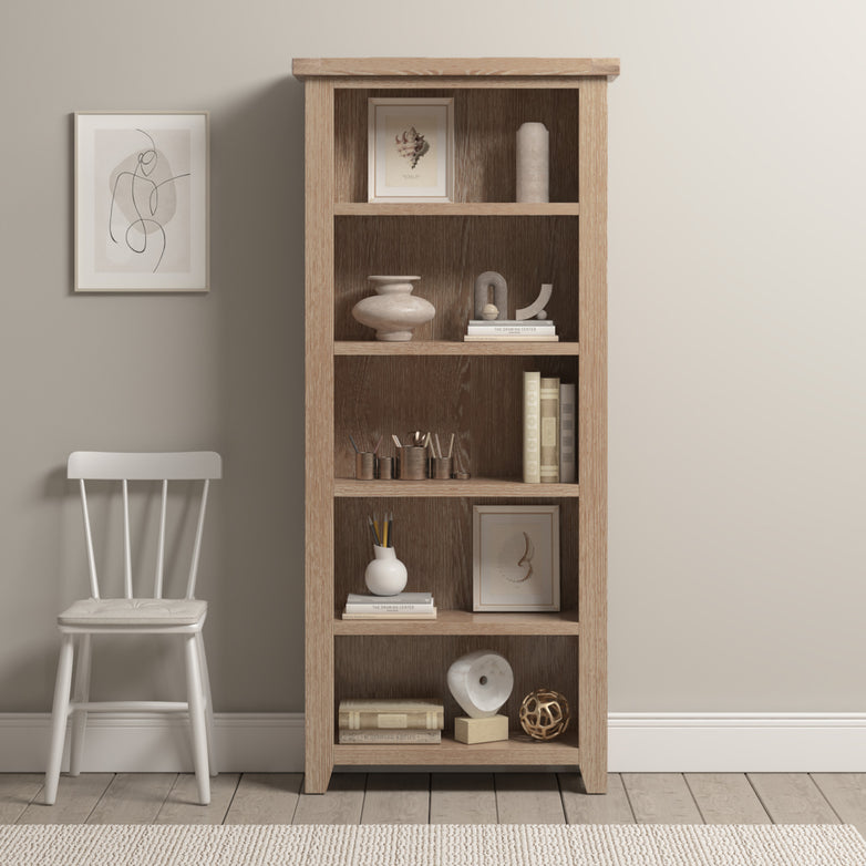 The Burford Large Bookcase in Natural Oak, decorated with art and decor items, offers versatile storage beside a white chair on a light rug over the tiled floor.