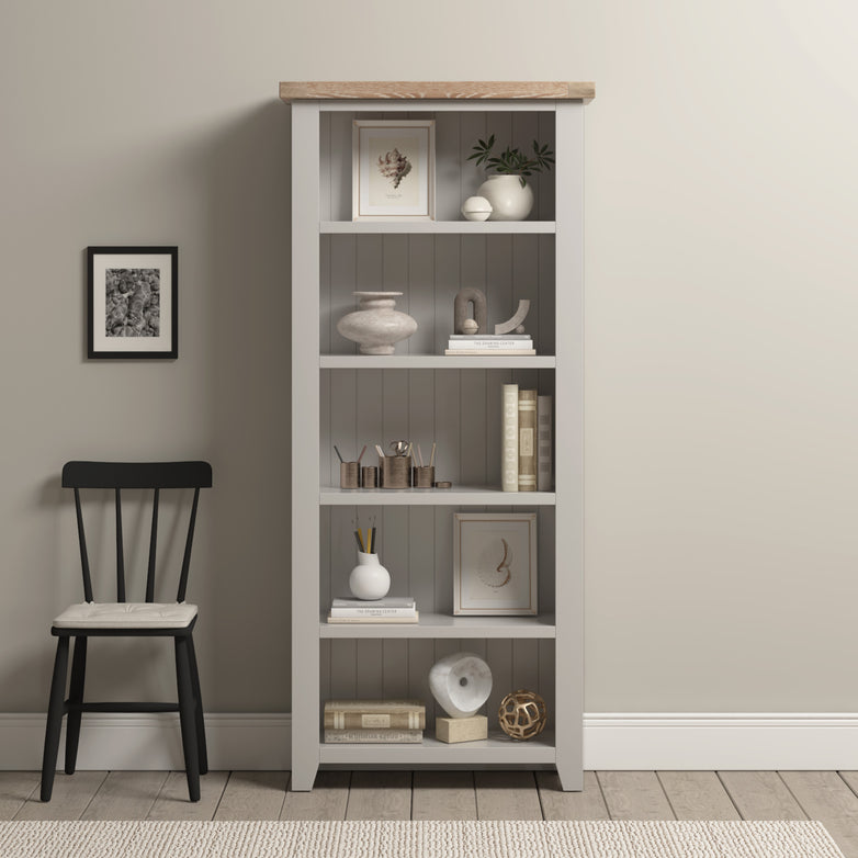 The Burford Large Bookcase in Pebble Grey showcases elegant decor and books, complemented by a black chair on a light rug in this minimalist room.