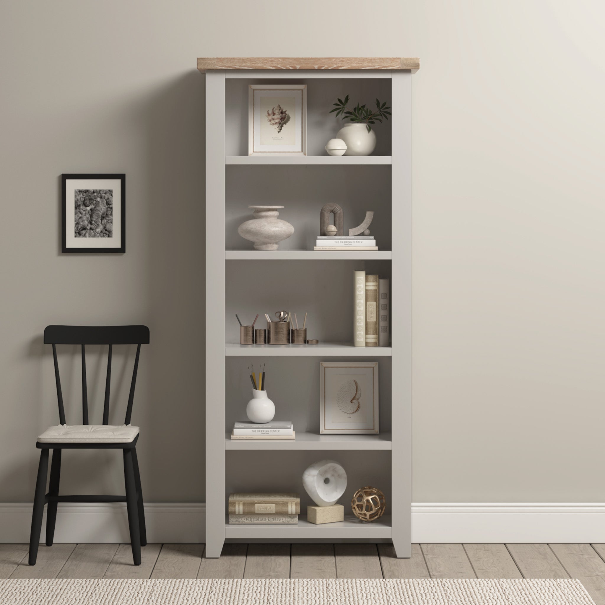 The Burford Large Bookcase in Pebble Grey, featuring an oak top, adds elegance with ample shelving for decor and books. A cozy chair nearby invites relaxation, while a small framed picture enhances the charm of the beige wall.