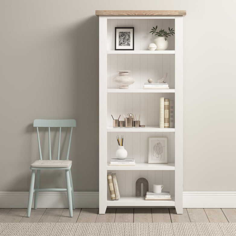 The Burford Large Bookcase in warm white showcases ample shelving space, adorned with decor and books, next to a light blue wooden chair on a beige carpeted floor.