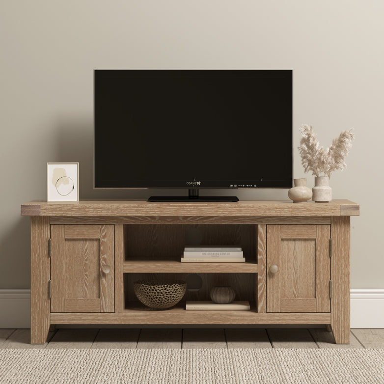 The Burford 120cm Media Unit in Natural Oak showcases a flat-screen TV, decor items, and a woven basket situated on a carpeted floor.