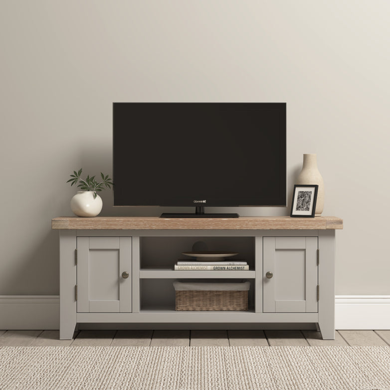 The Burford 120cm Media Unit in Pebble Grey features an elegant wooden stand with cabinets and shelves, showcasing a TV adorned with a plant, framed photo, and vase, perfectly enhancing the minimal room's aesthetic.