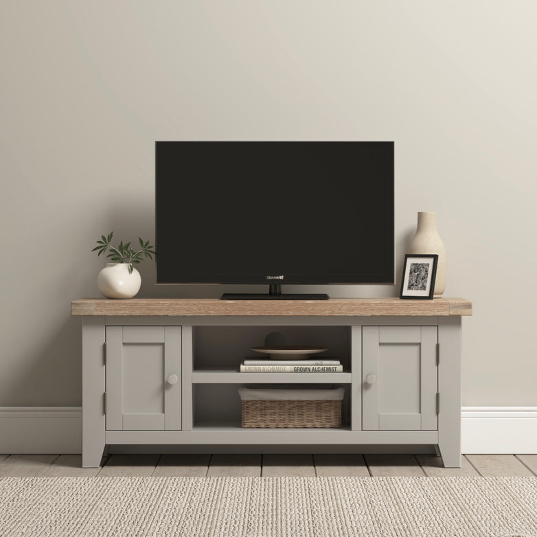 A Burford 120cm Media Unit in pebble grey displays a TV on its stylish wooden stand with sleek cabinets. Accentuated by a vase, framed photo, and small plant, it provides an elegant contrast against the natural oak wall.