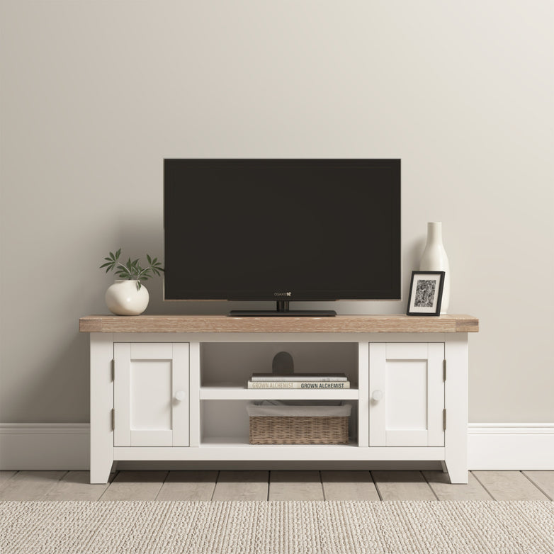 The Burford 120cm Media Unit in warm white has a wooden top displaying a TV, vase, framed photo, and plant, with a basket neatly placed on the open shelf below.