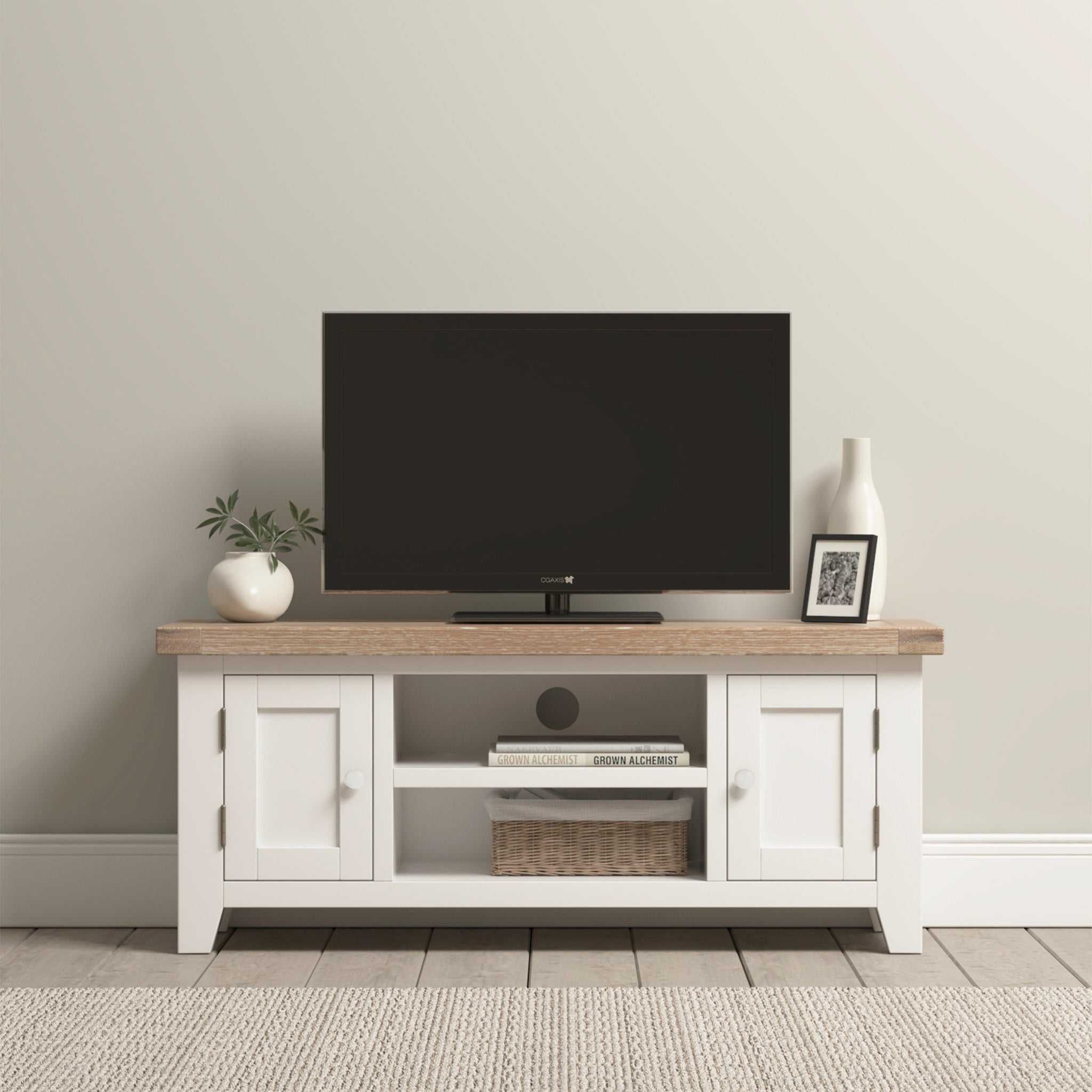 TV on a Burford 120cm Media Unit in Warm White, showcasing its rustic charm with a white-washed oak top. It stands elegantly next to white cabinets, adorned with a plant, vase, and framed photo. The neutral wall and rug provide an inviting and serene backdrop.