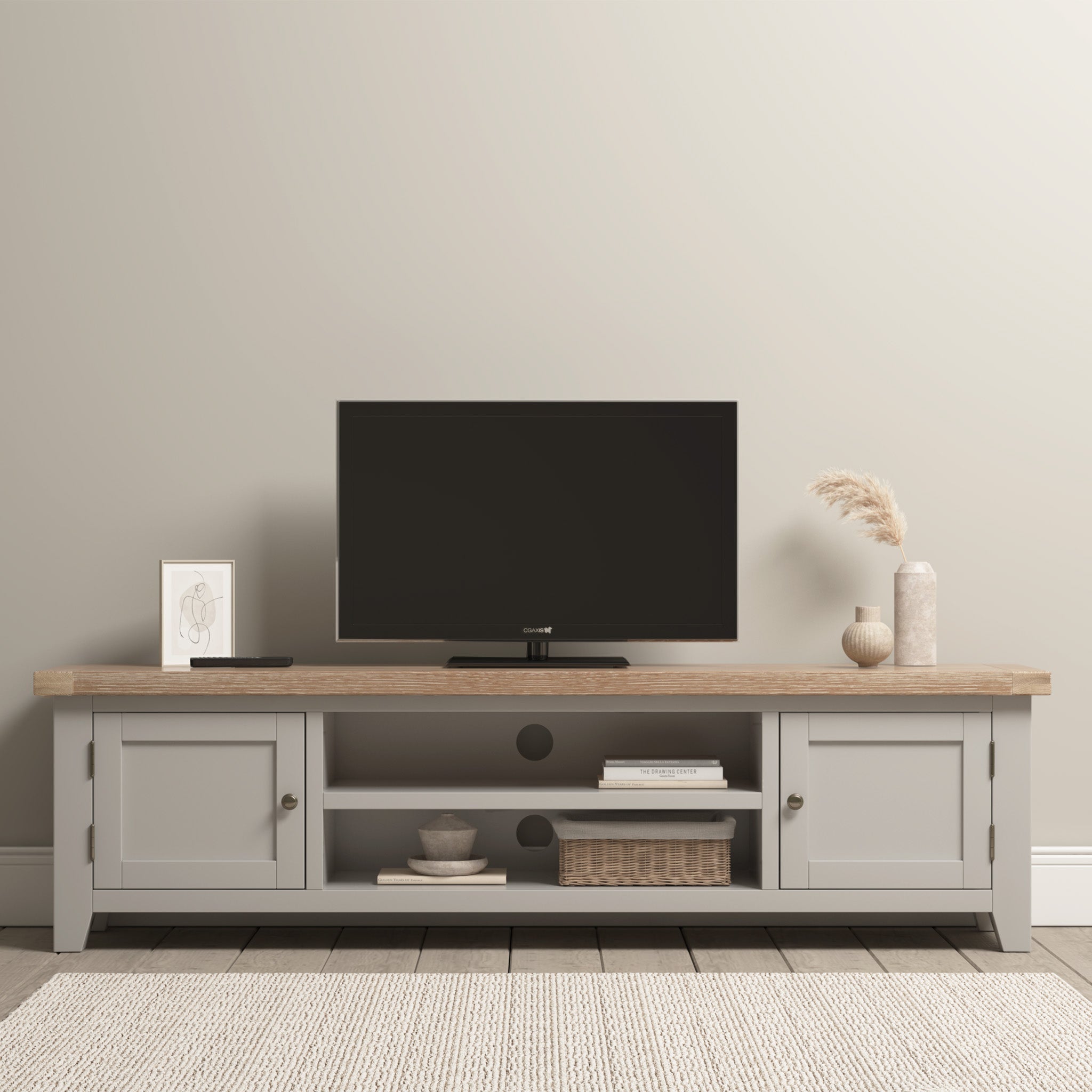 A minimalist living room showcasing a TV on the Burford Large 180cm Media Unit in Pebble Grey, adorned with decorative items and a textured rug. The space is tastefully complemented by a pair of ladderback dining chairs in natural oak with fabric seats, adding warmth and elegance to the decor.