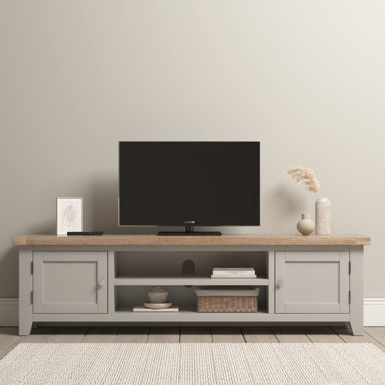 The Burford Large 180cm Media Unit in Pebble Grey holds a small TV, framed art, vases, and a basket. Its neutral tones and the soft-light rug complement a nearby ladderback dining chair in natural oak, enhancing the elegant arrangement.