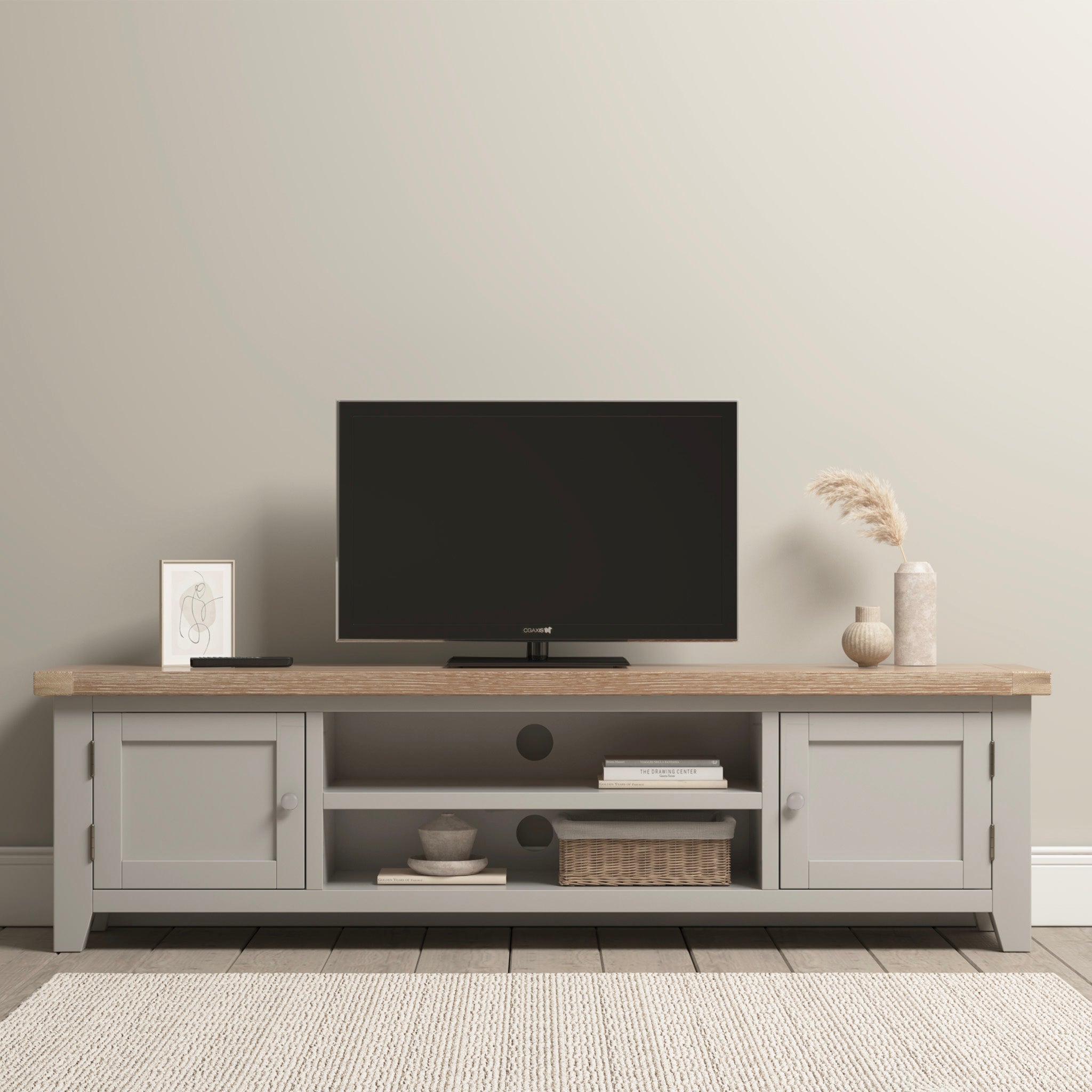 A minimalist living room showcases a TV atop the Burford Large 180cm Media Unit in Pebble Grey, paired with subtle decor items against beige walls. A cozy fabric seat and the classic Burford Ladderback Dining Chair enhance the room's charm.