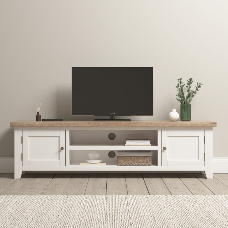 A minimalist living room showcases a TV on the Burford Large 180cm Media Unit in Warm White, accented with decor items and a neutral rug. A Burford Ladderback Dining Chair with a fabric seat adds charm, enhancing the natural oak hues subtly present in the serene space.