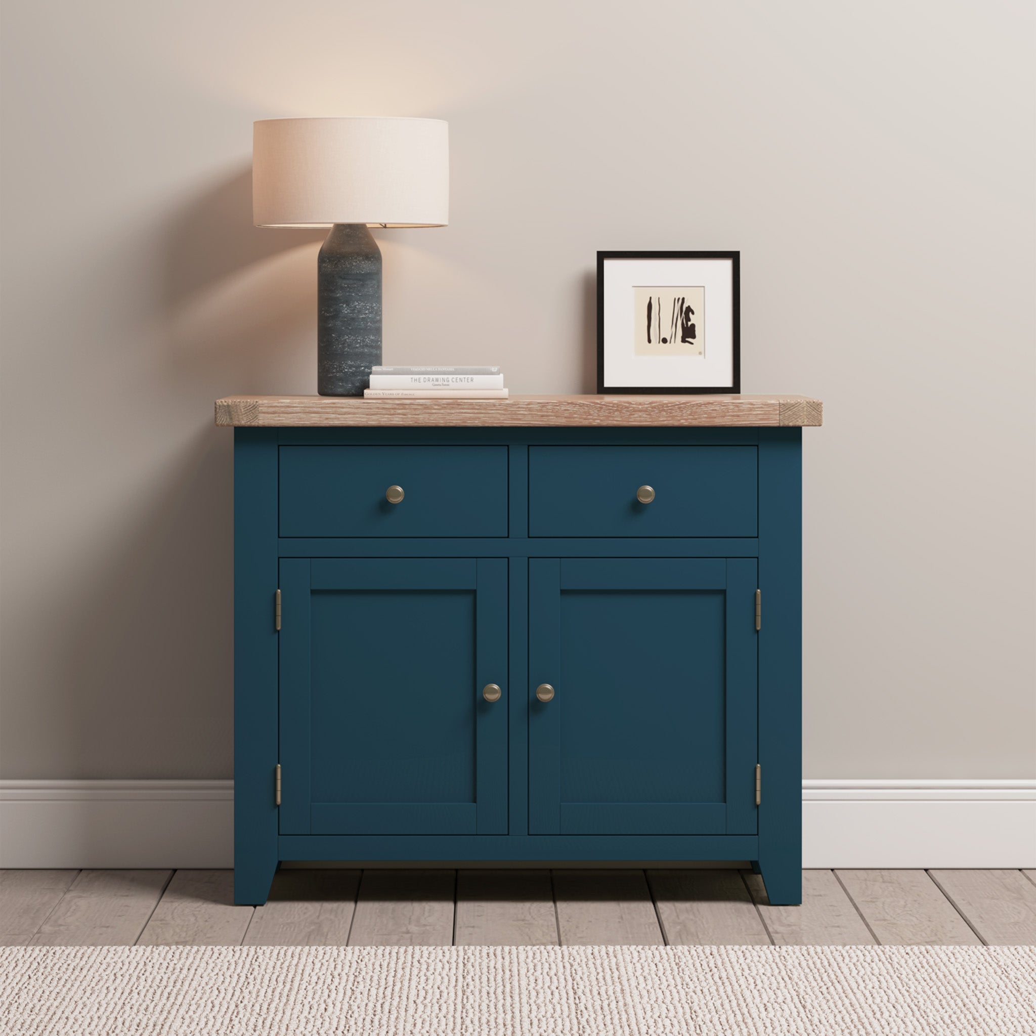 The Burford 2 Door 2 Drawers Sideboard in navy blue features a natural oak top, beautifully adorned with a lamp, books, and framed art, set against a light-colored wall.