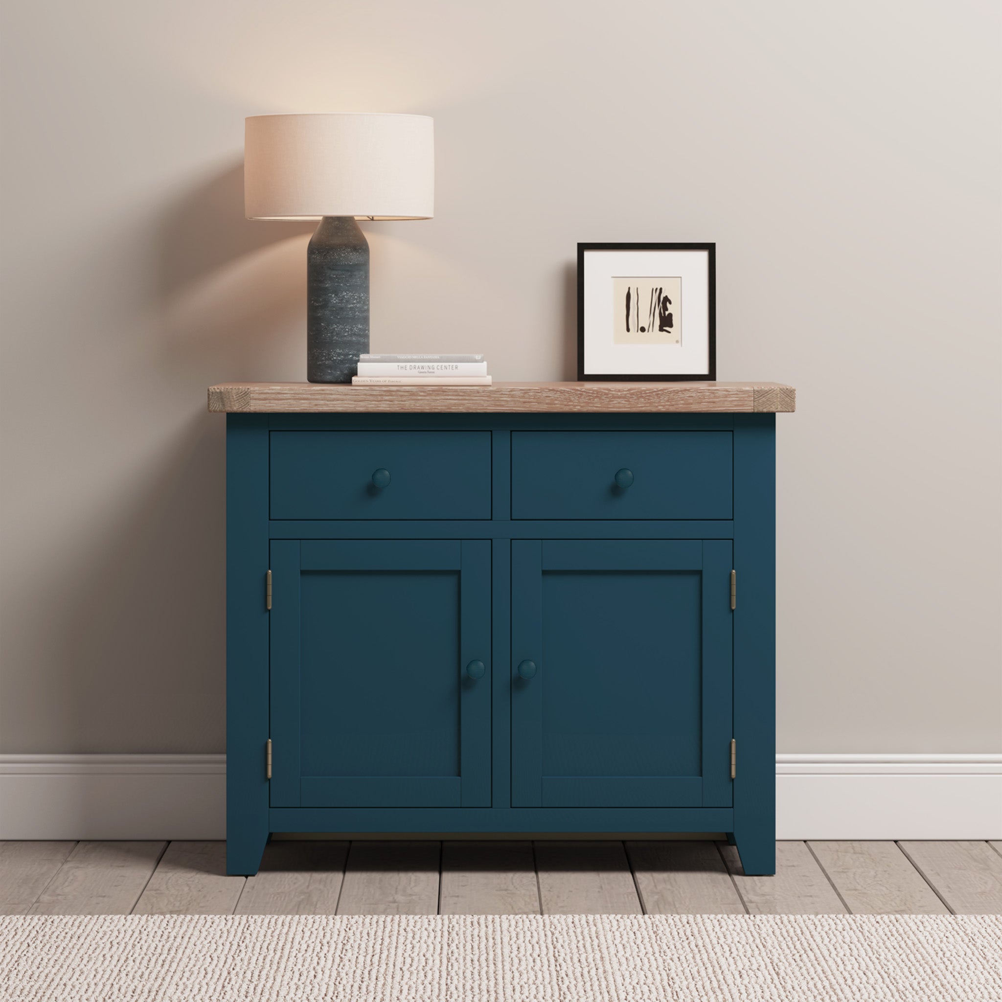 The Burford 2 Door 2 Drawers Sideboard in Navy Blue, decorated with a lamp, books, and framed art, stands elegantly against a light wall and tile floor.