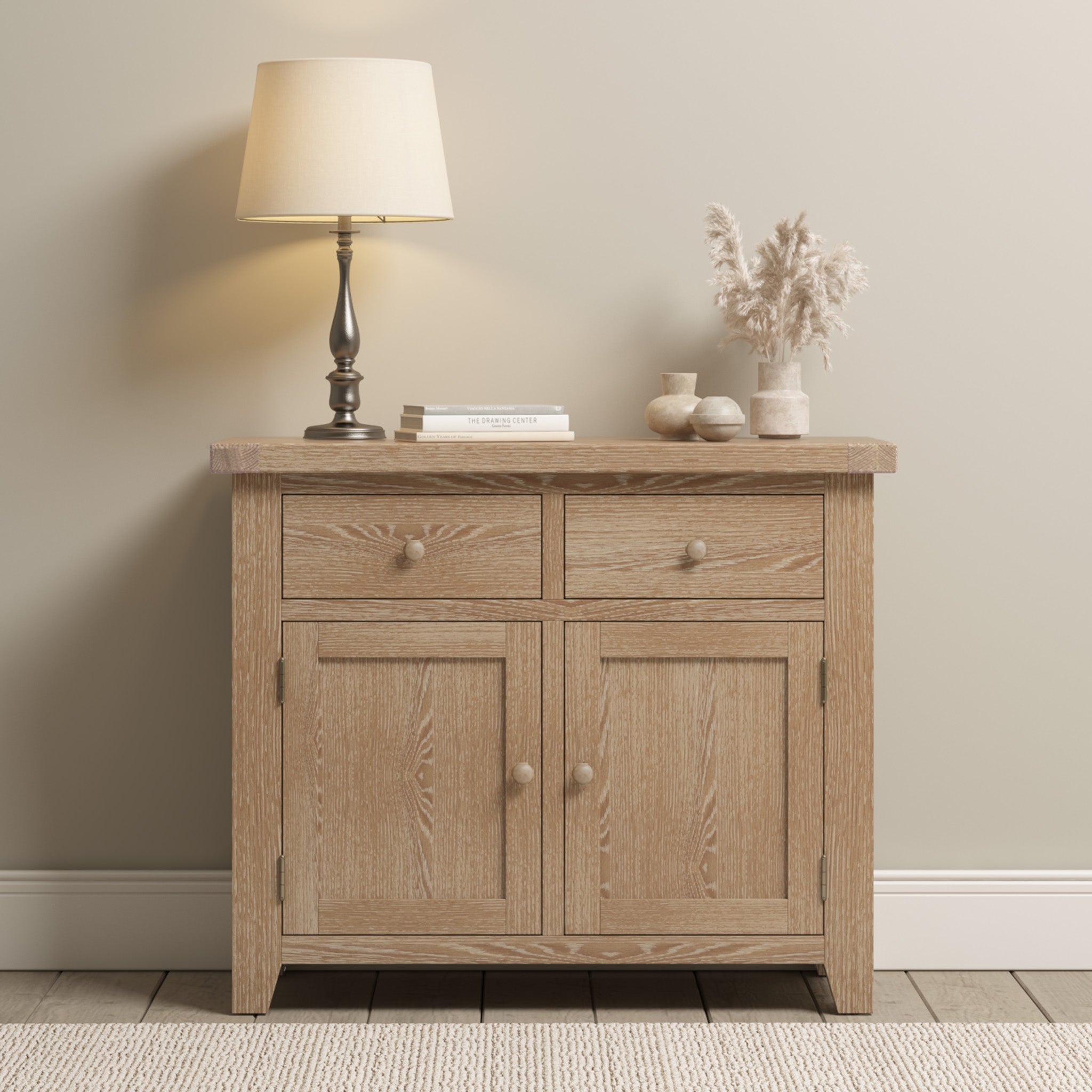 The Burford 2 Door 2 Drawers Sideboard in Natural Oak brings a touch of rustic charm to a neutral-toned room, beautifully complemented by a lamp, books, and ceramic decor.