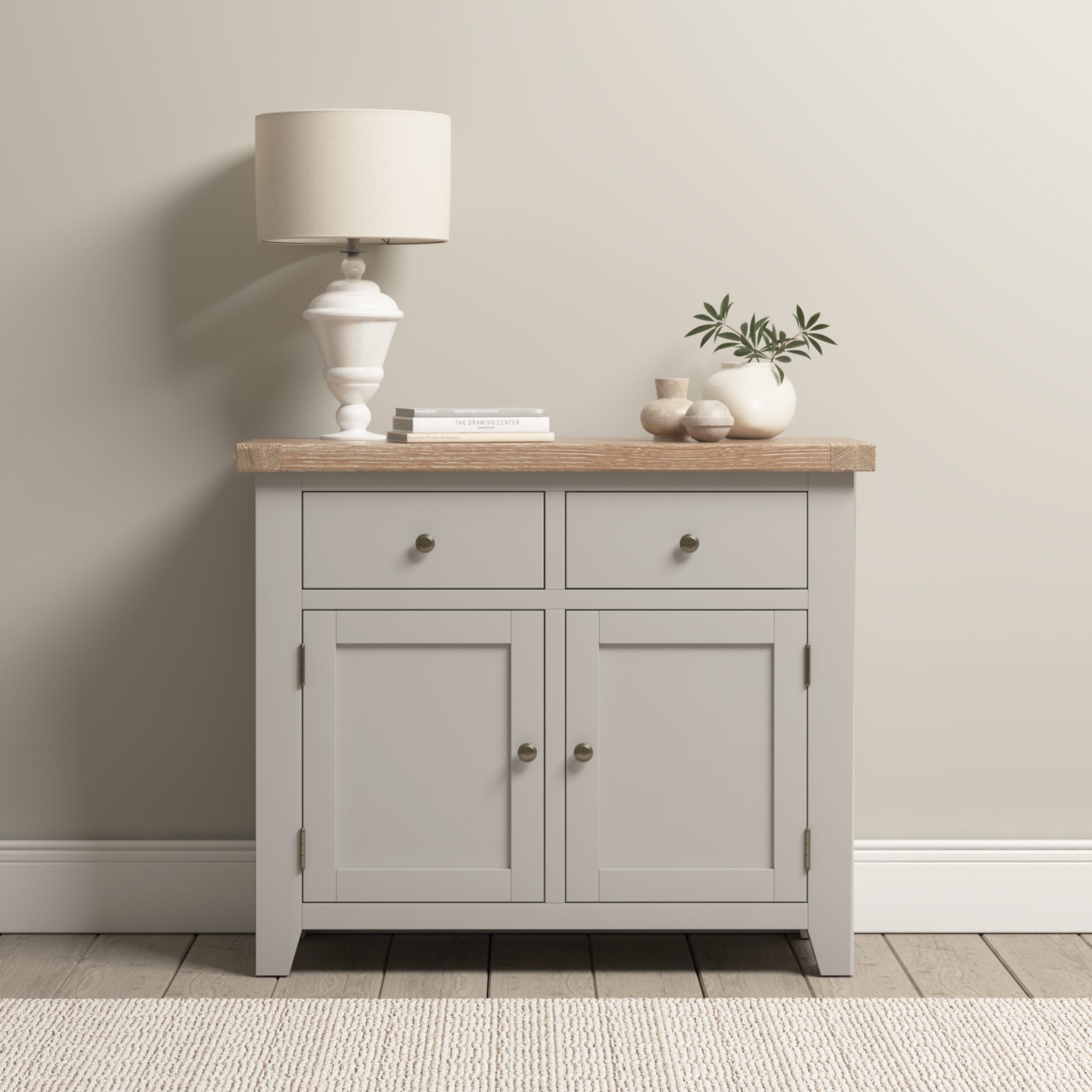 The Burford 2 Door 2 Drawers Sideboard in Pebble Grey, featuring a natural oak top, is adorned with a lamp, vase, books, and plant. It rests elegantly against a beige wall on a hardwood floor.