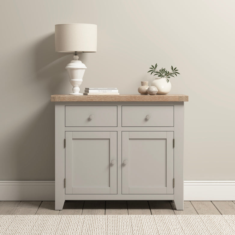 A Burford 2 Door 2 Drawers Sideboard in Pebble Grey elegantly displays a lamp, books, and decor beneath a subtle beige wall, complemented by natural oak accents and a chic vase on top.