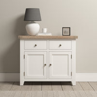 The Burford 2 Door 2 Drawers Sideboard in Warm White showcases a sleek white cabinet with two drawers and doors. On top of the sideboard, a gray lamp, a book, a mug, and a photo frame are gracefully arranged against a light-colored wall backdrop.