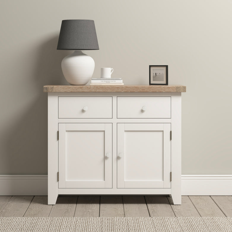 The Burford 2 Door 2 Drawers Sideboard in Warm White, featuring a wooden top, elegantly displays a lamp, books, a cup, and a framed photo against the backdrop of an oak floor.