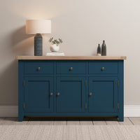 The Burford Large 3 Door 3 Drawers Sideboard in Navy Blue, with its elegant wooden top, displays a lamp, books, a plant, and vases against a neutral wall.