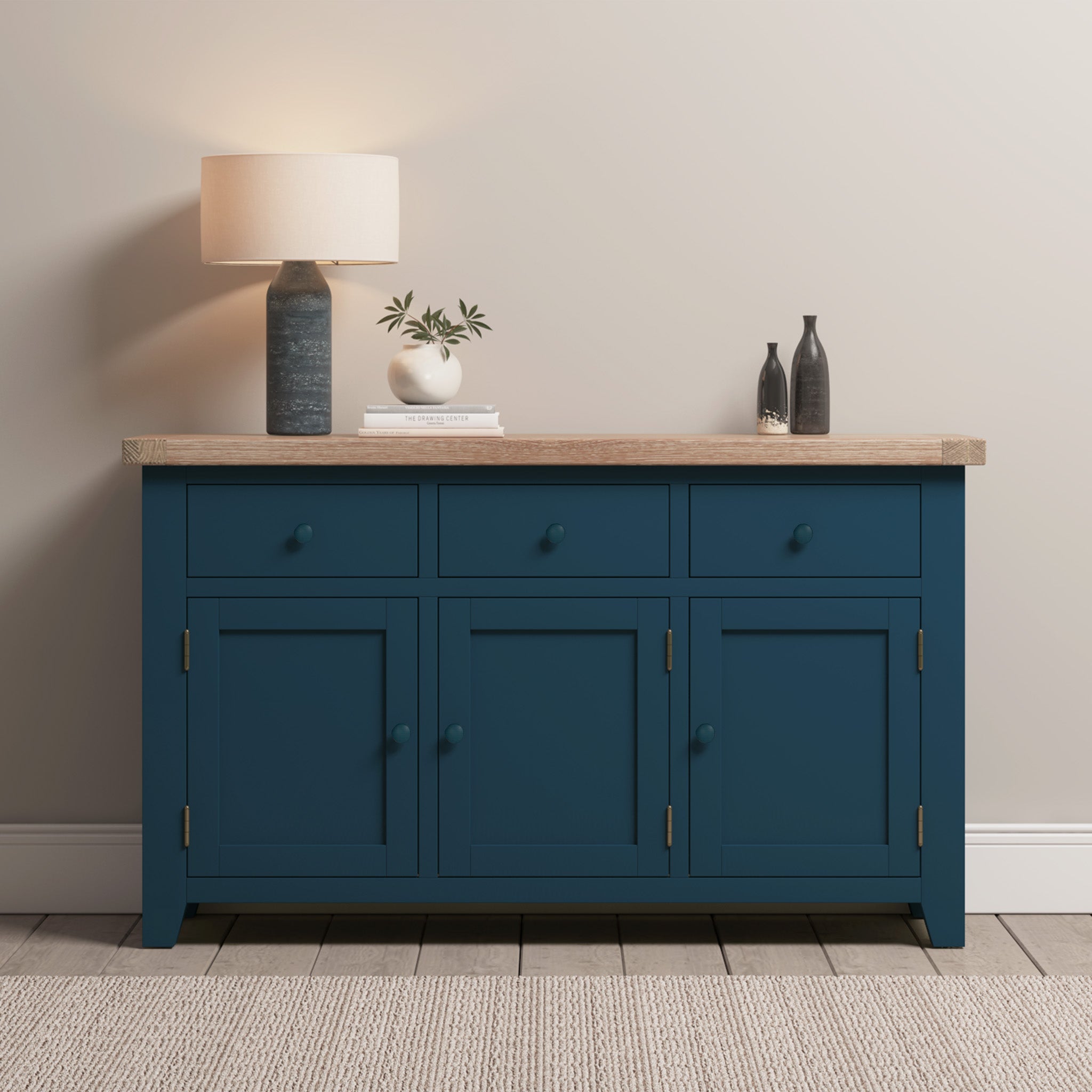 The Burford Large 3 Door 3 Drawers Sideboard in navy blue showcases a premium natural oak top, elegantly decorated with a lamp, vases, and books. It sits beautifully against a neutral wall over a light rug.