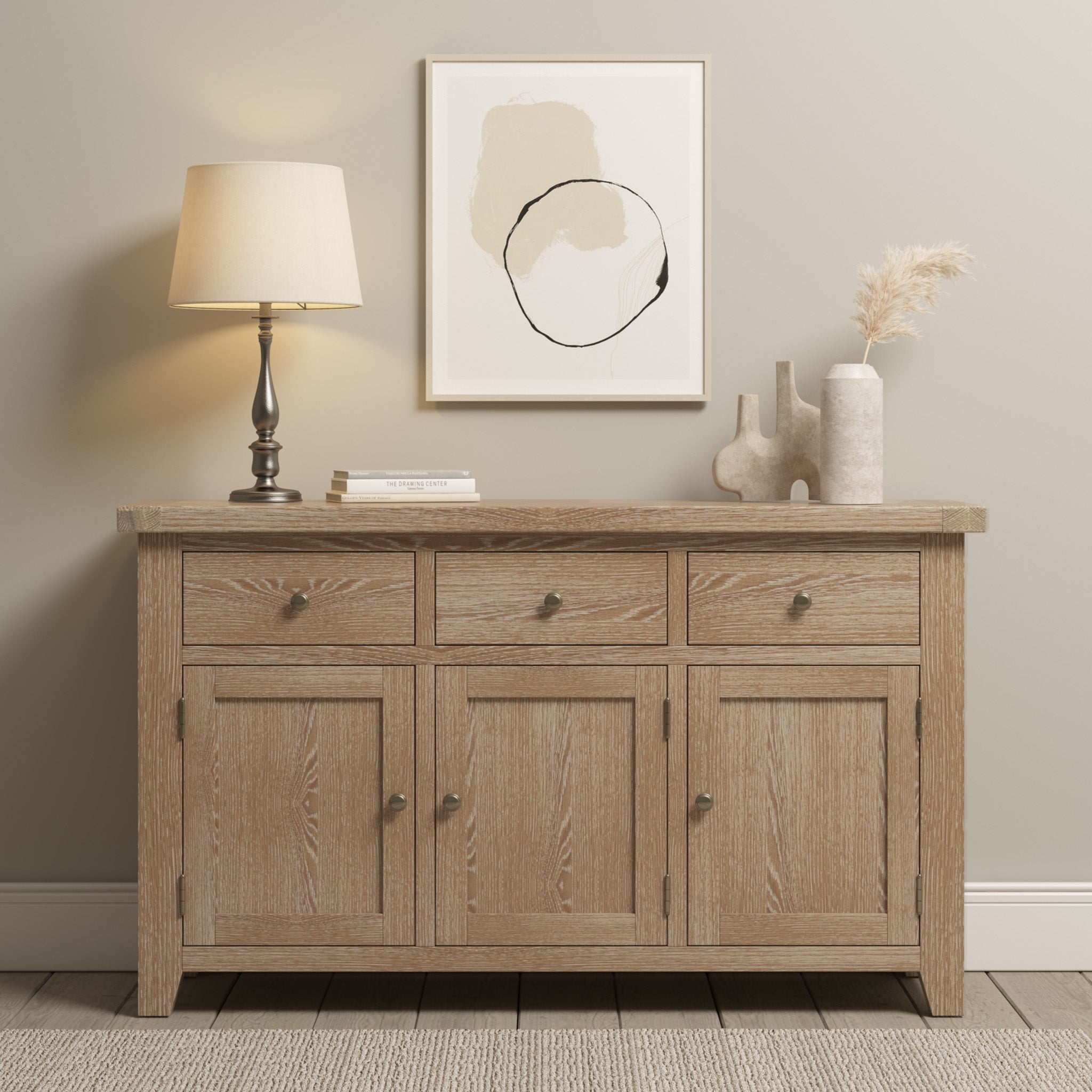 The Burford Large 3 Door 3 Drawers Sideboard in Natural Oak, crafted with elegance, features three doors and three drawers. It beautifully complements the lamp, books, vases, and abstract art against a light-colored wall. A beige rug lies beneath.