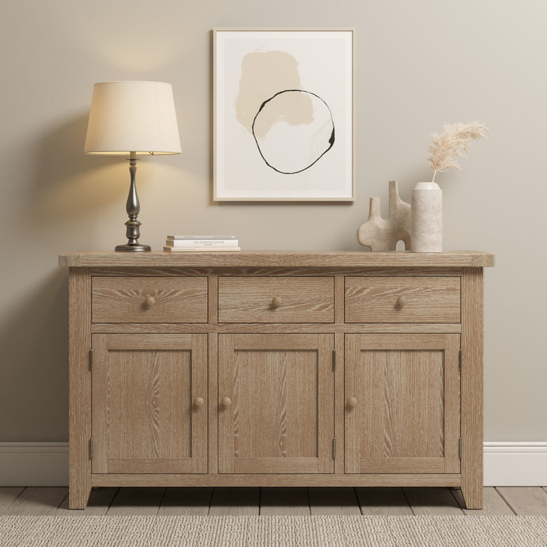 The Burford Large 3 Door 3 Drawers Sideboard in Natural Oak boasts a design that includes drawers and gracefully supports a lamp, abstract art, and decorative vases. With its placement against a neutral wall and rug background, this piece seamlessly elevates any room's decor.