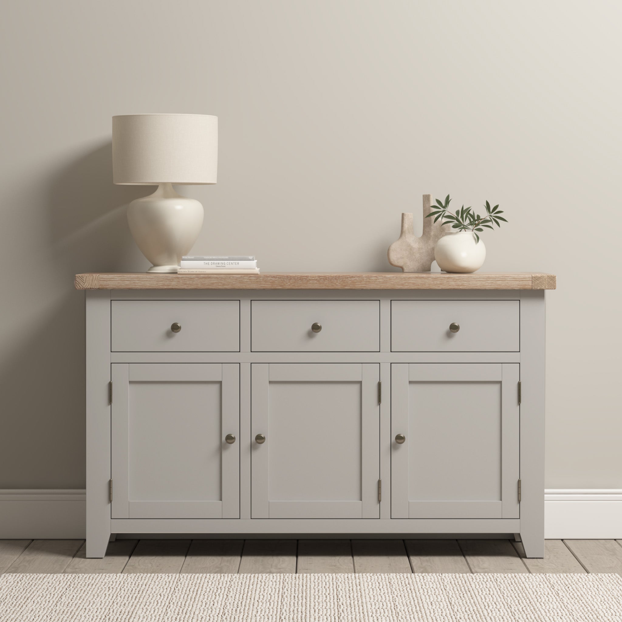 The Burford Large 3 Door 3 Drawers Sideboard in Pebble Grey showcases a natural oak top, beautifully decorated with a lamp, book, and ceramics, and is placed against a beige wall.