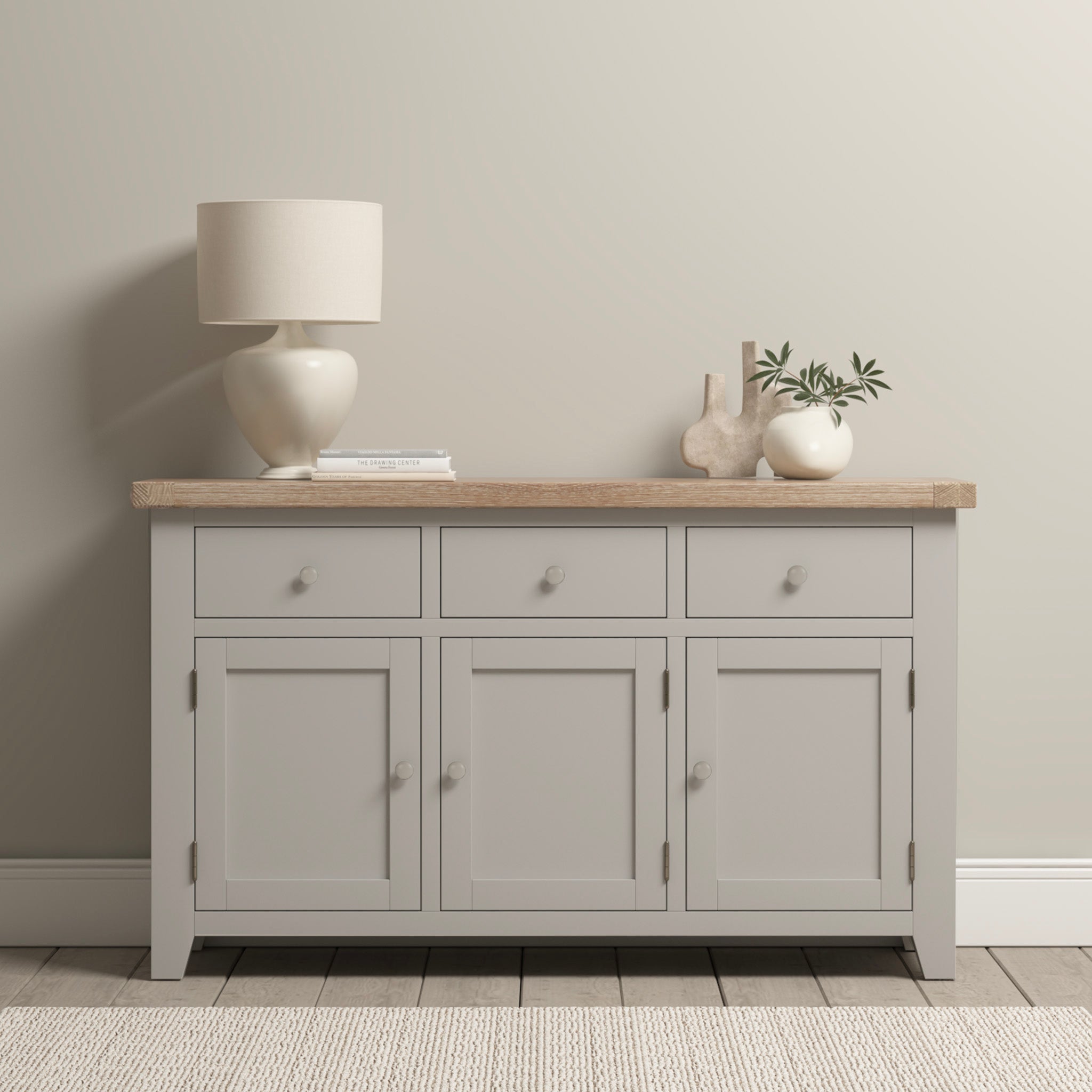 A Burford Large 3 Door 3 Drawers Sideboard in Pebble Grey with a natural oak top, adorned with a lamp, books, and decorative vases, rests elegantly against a neutral wall.