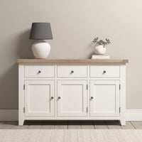 The Burford Large 3 Door 3 Drawers Sideboard in Warm White, featuring a natural oak top, elegantly displays a lamp and a small potted plant against a neutral wall, providing both style and storage.