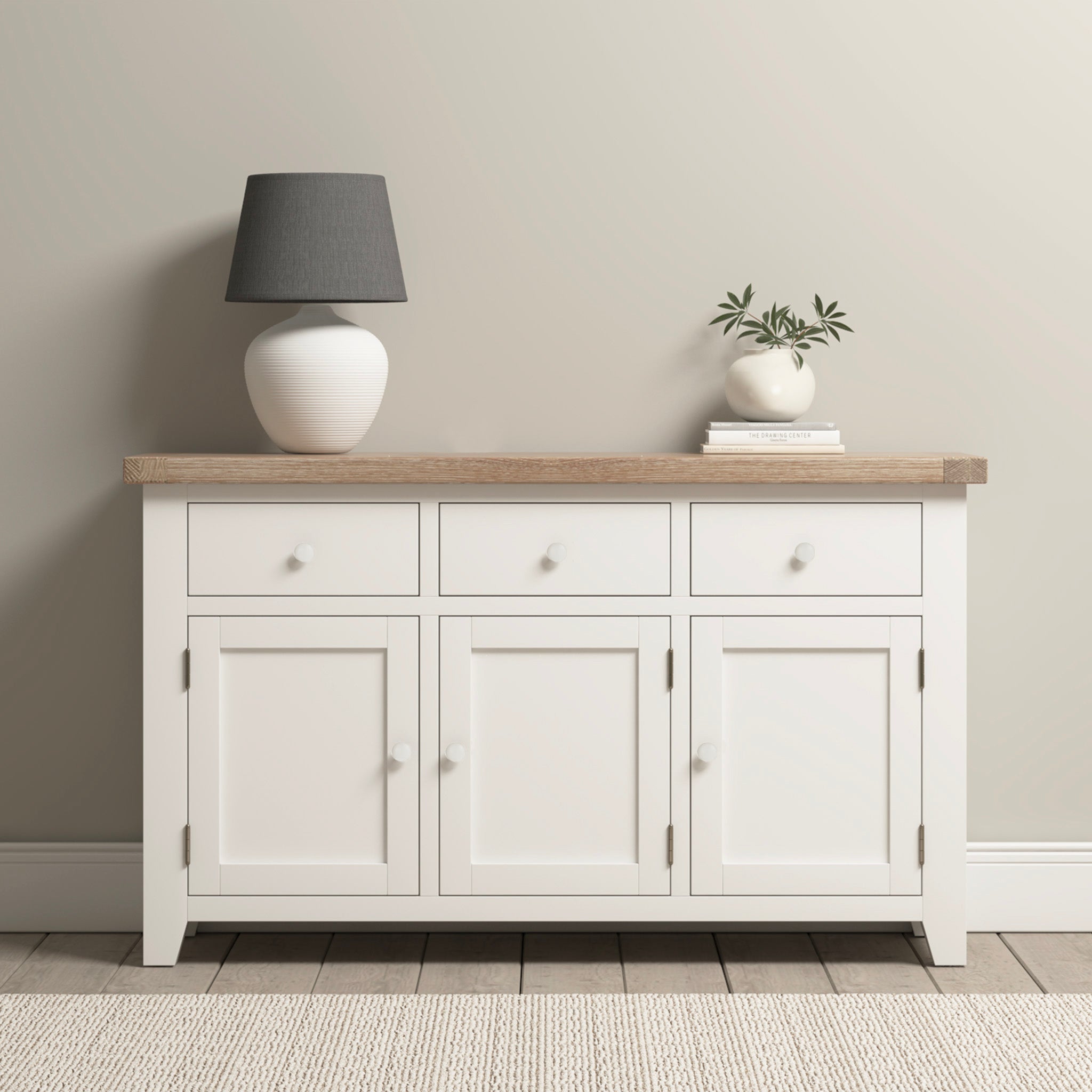 The Burford Large 3 Door 3 Drawers Sideboard in warm white stands elegantly against a neutral wall, adorned with a lamp and potted plant on top, perfectly complementing the carpeted floor while providing ample storage.