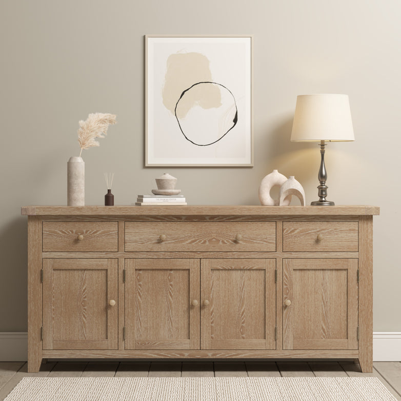 Burford Large 4 Door 3 Drawers Sideboard in Natural Oak adorned with decor, a lamp, and abstract art graces the wall in a neutral-toned room.