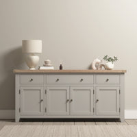 The Burford Large 4 Door 3 Drawers Sideboard in Pebble Grey offers spacious storage with its multiple drawers, elegantly topped with a lamp, decor items, and a potted plant, creating a contemporary look against a light wall.