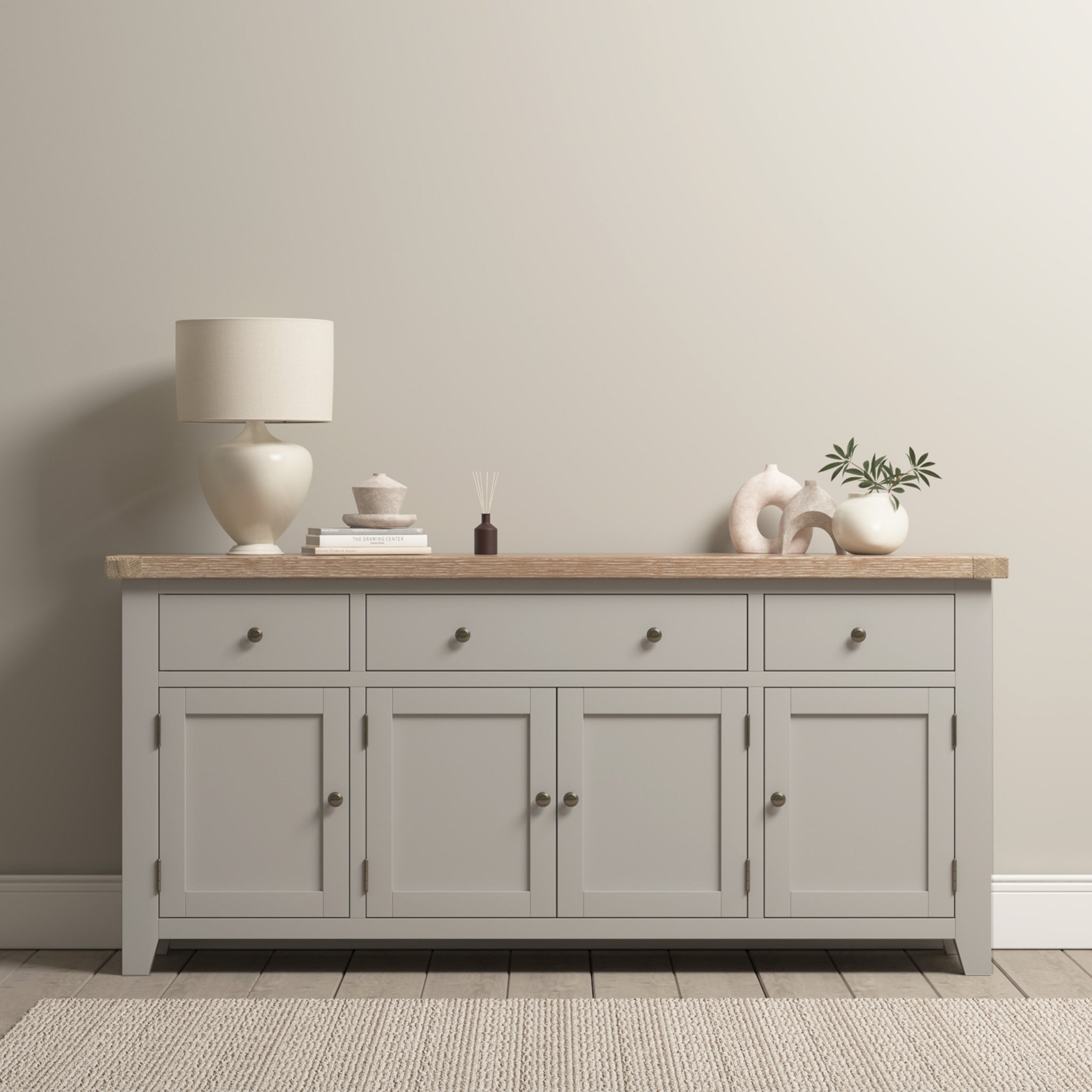 The Burford Large 4 Door 3 Drawers Sideboard in Pebble Grey offers spacious storage with its multiple drawers, elegantly topped with a lamp, decor items, and a potted plant, creating a contemporary look against a light wall.