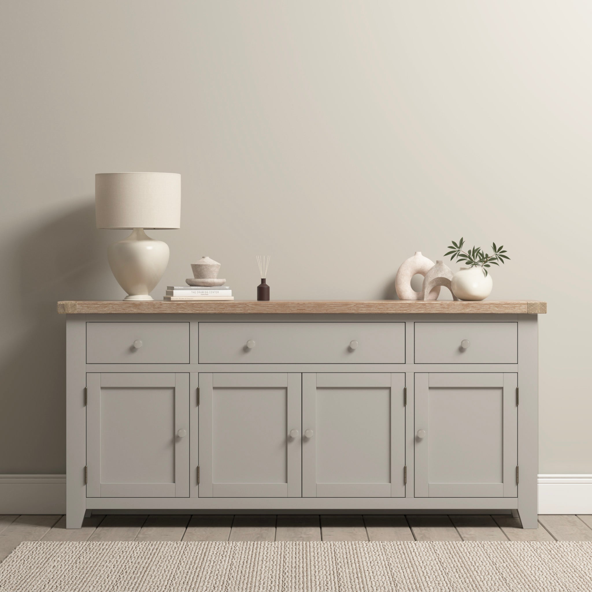 The Burford Large 4 Door 3 Drawers Sideboard in Pebble Grey exudes contemporary elegance, with a lamp, books, decor, and plant artfully arranged against a neutral-colored wall. Its spacious storage ensures style meets function effortlessly.