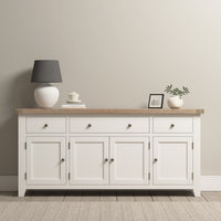 Burford Large 4 Door 3 Drawers Sideboard in Warm White with a natural oak top, table lamp, small plant, framed photo, and books against a light gray wall provides ample storage space.