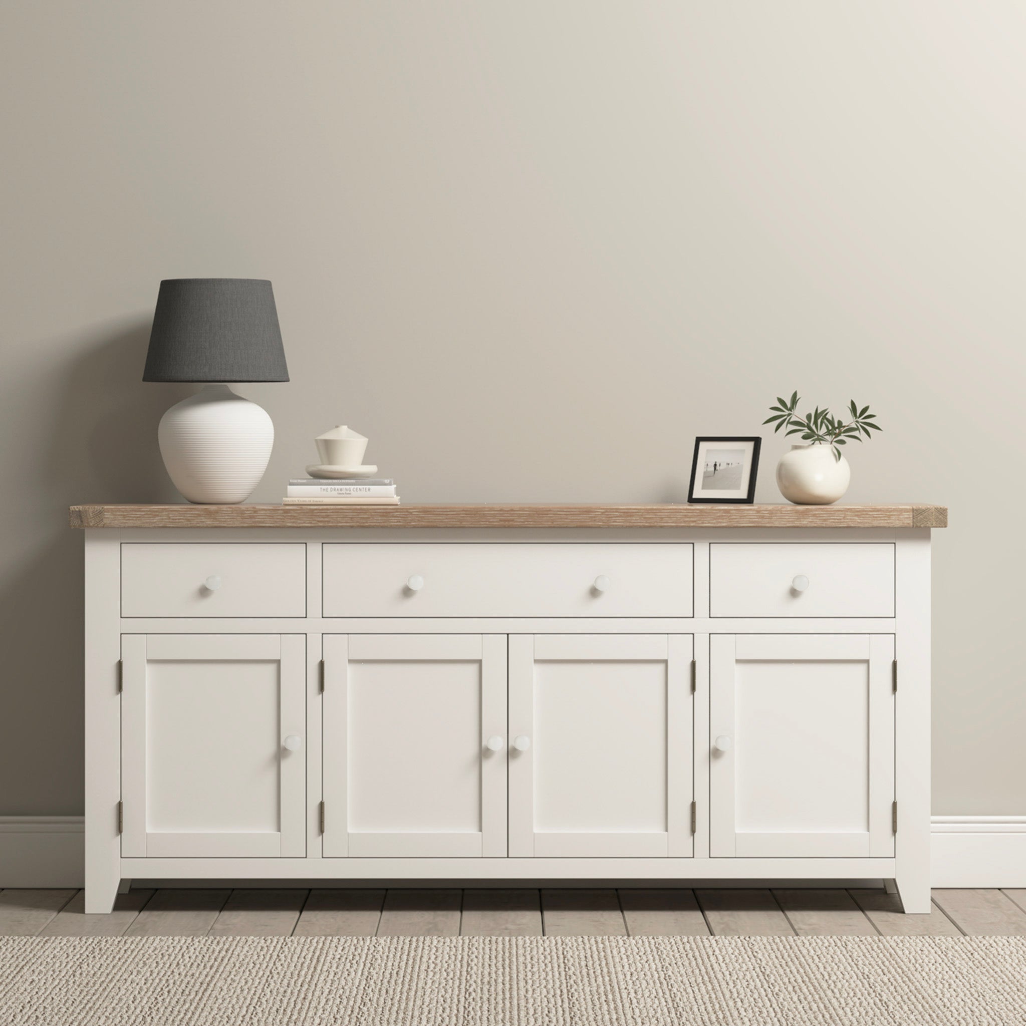 The Burford Large 4 Door 3 Drawers Sideboard in Warm White showcases a natural oak top adorned with a lamp, framed photo, and small potted plant against a beige wall, providing stylish storage.