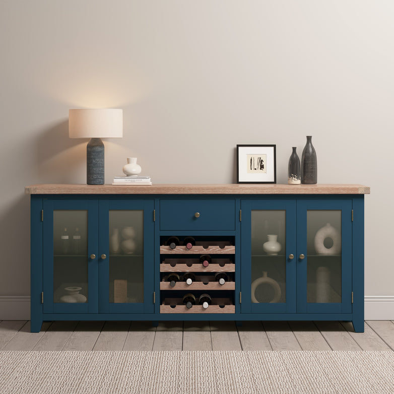 The Burford Grand Sideboard with Wine Rack in Navy Blue showcases a wine rack and glass doors and is elegantly topped with a stylish lamp, with vases and art enhancing its presence against the light wall.