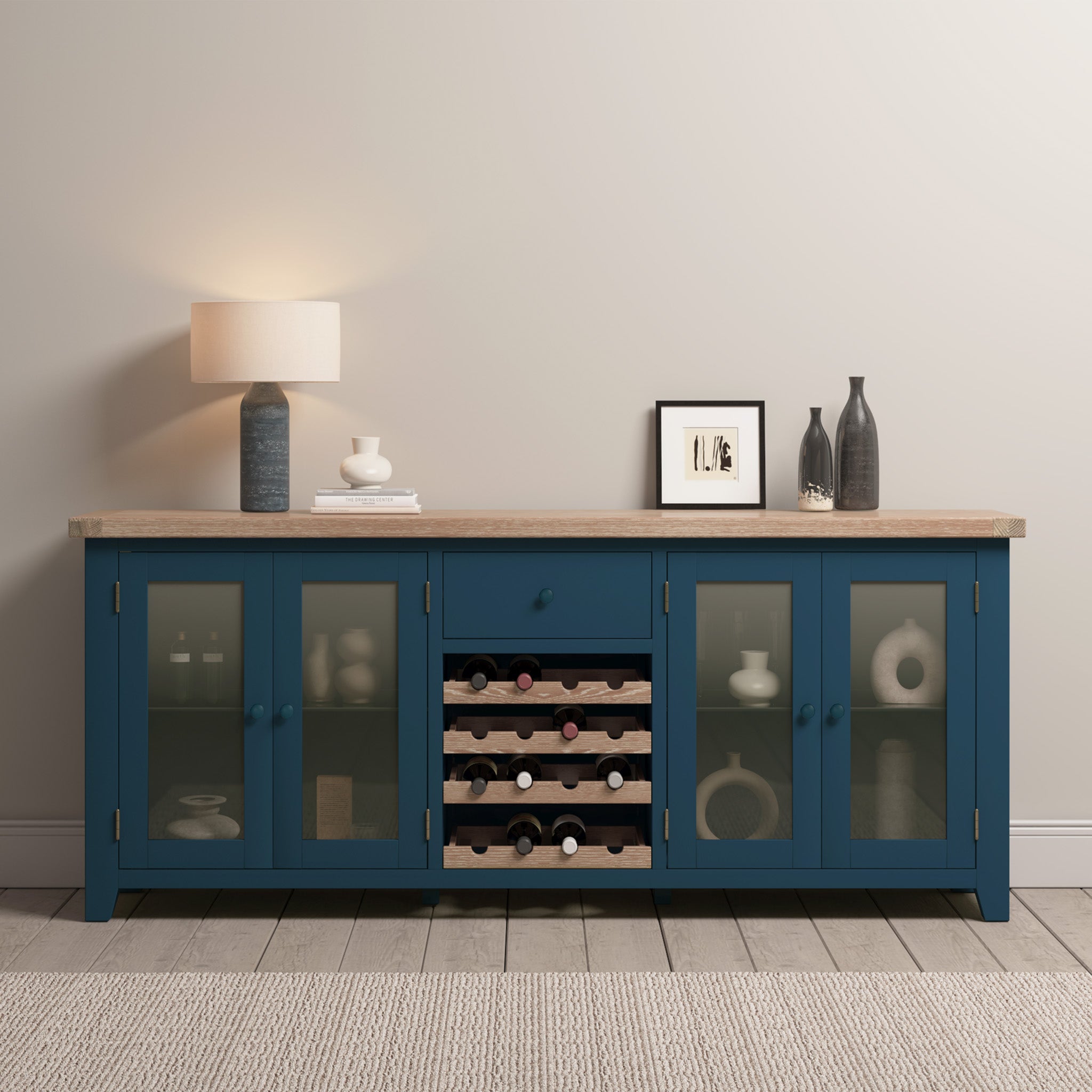 The Burford Grand Sideboard with Wine Rack in Navy Blue showcases sophisticated glass doors and a built-in oak wine rack, adorned with a lamp and decorative pieces, beautifully complementing the light-colored wall.