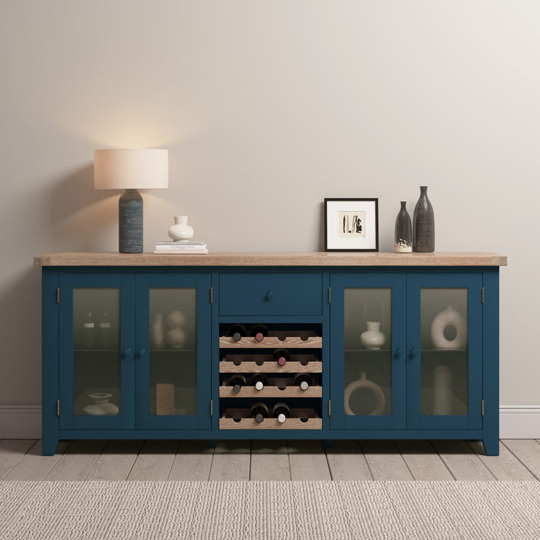 The Burford Grand Sideboard with Wine Rack in Navy Blue features natural oak accents, navy blue cabinets, and a built-in wine rack. It is tastefully decorated with a lamp and ornamental pieces, all set against a beige wall on a gray rug.